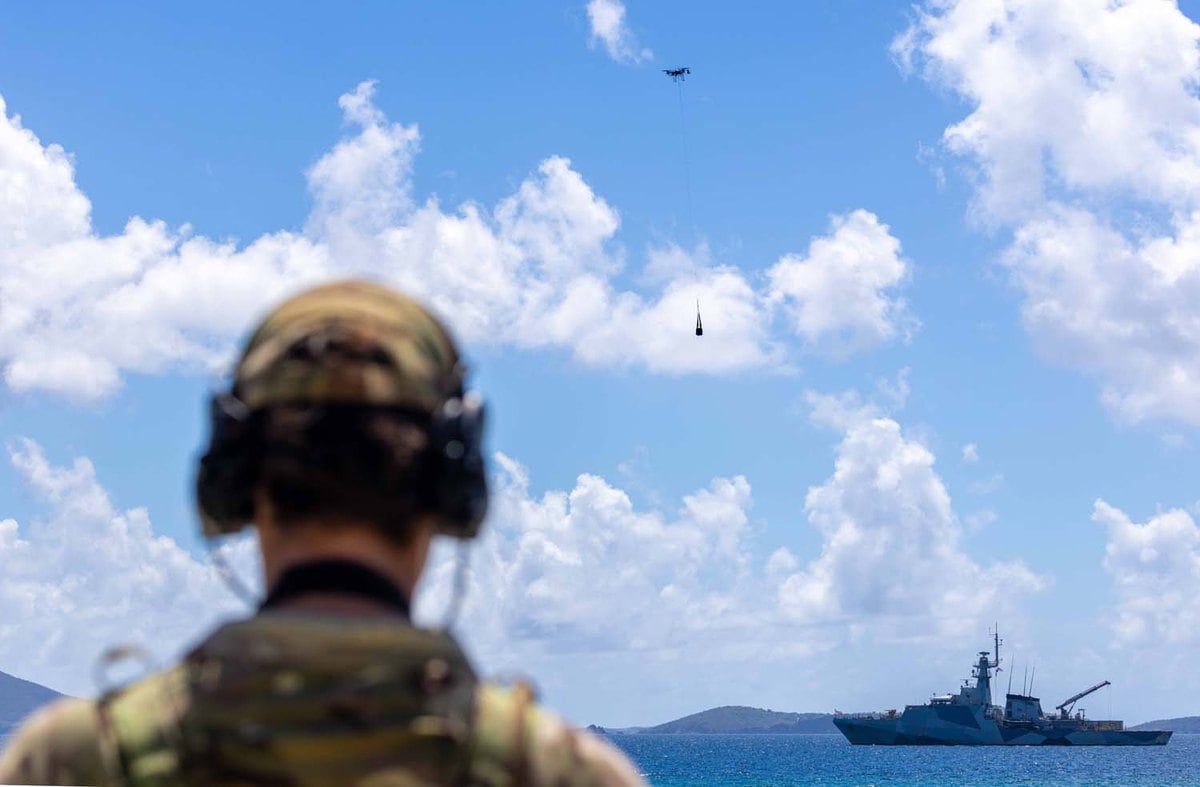 Un militar de espaldas observa un dron Malloy que transporta una carga colgada, con el HMS Trent al fondo en el mar