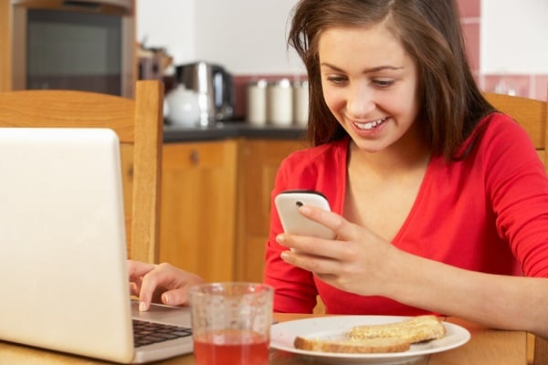 Teenage Girl Using Laptop And Mobile Phone Whilst Eating Breakfast