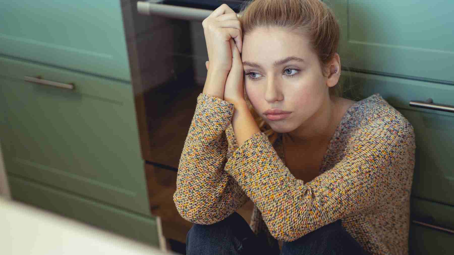 Young woman sitting on kitchen floor looking thoughtful, reflecting hesitation before adopting a new social norm