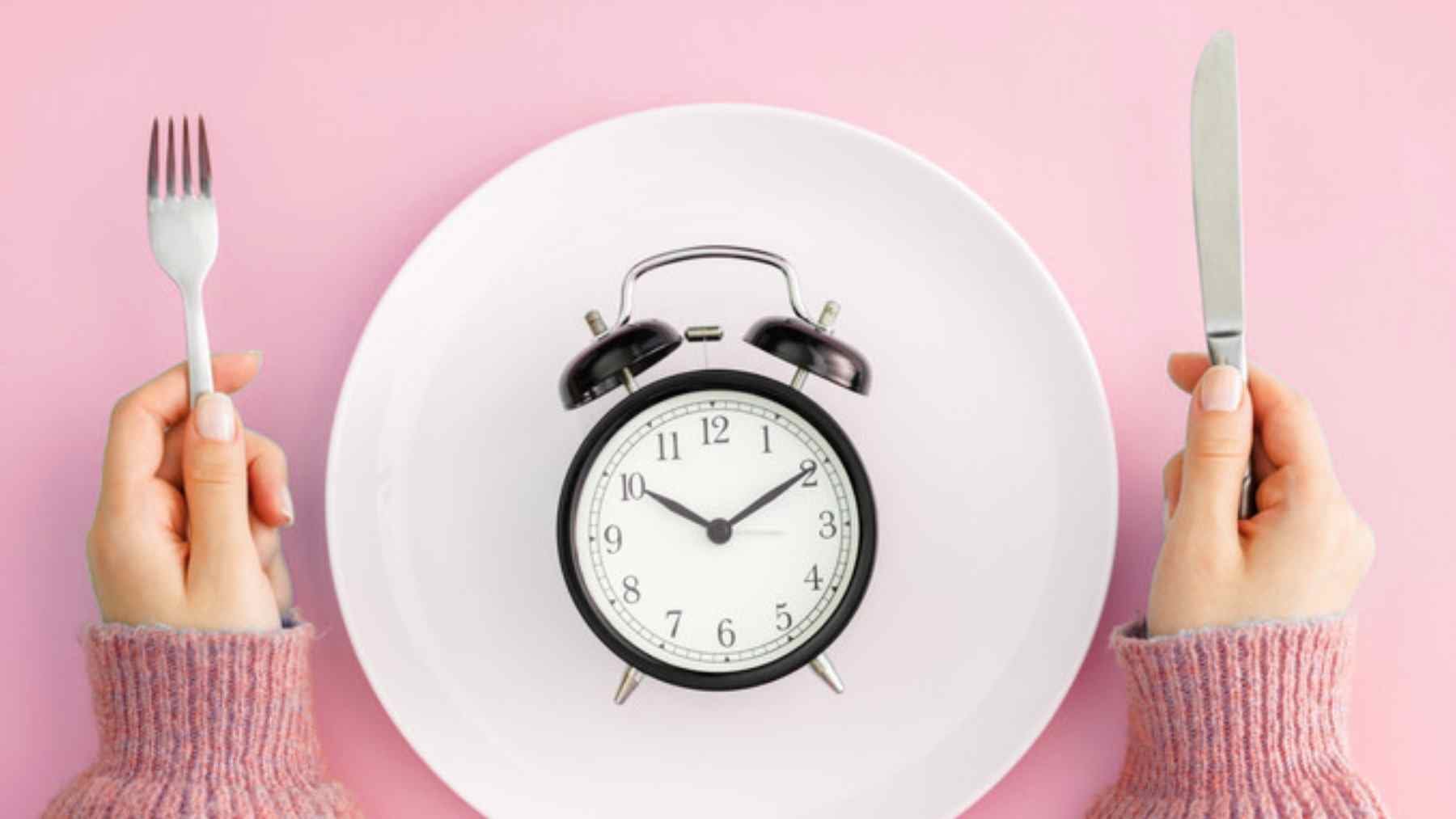 Intermittent fasting concept with a clock on a dinner plate and hands holding fork and knife