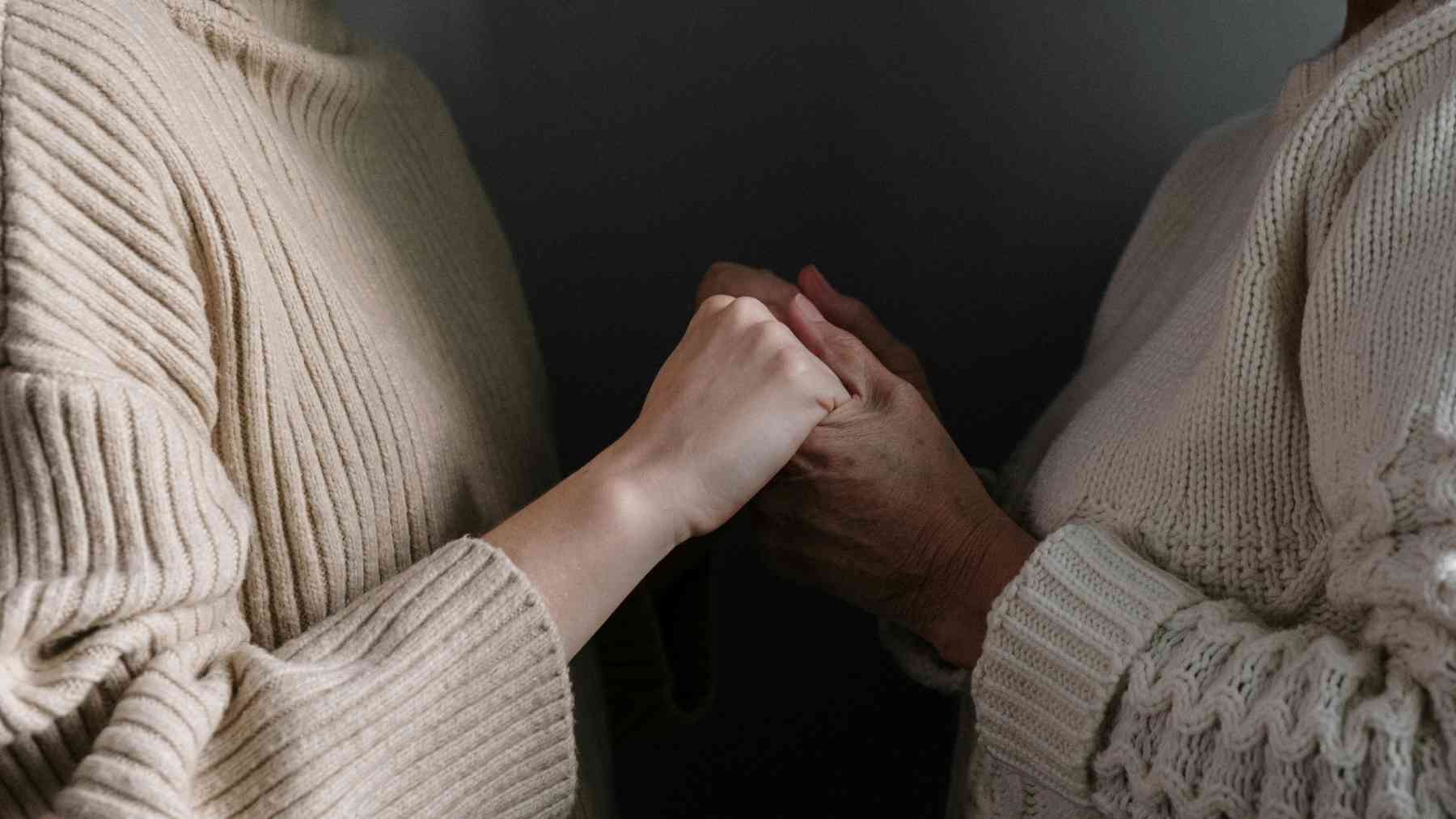 A younger person holds an older woman’s hands while listening with patience and emotional connection.