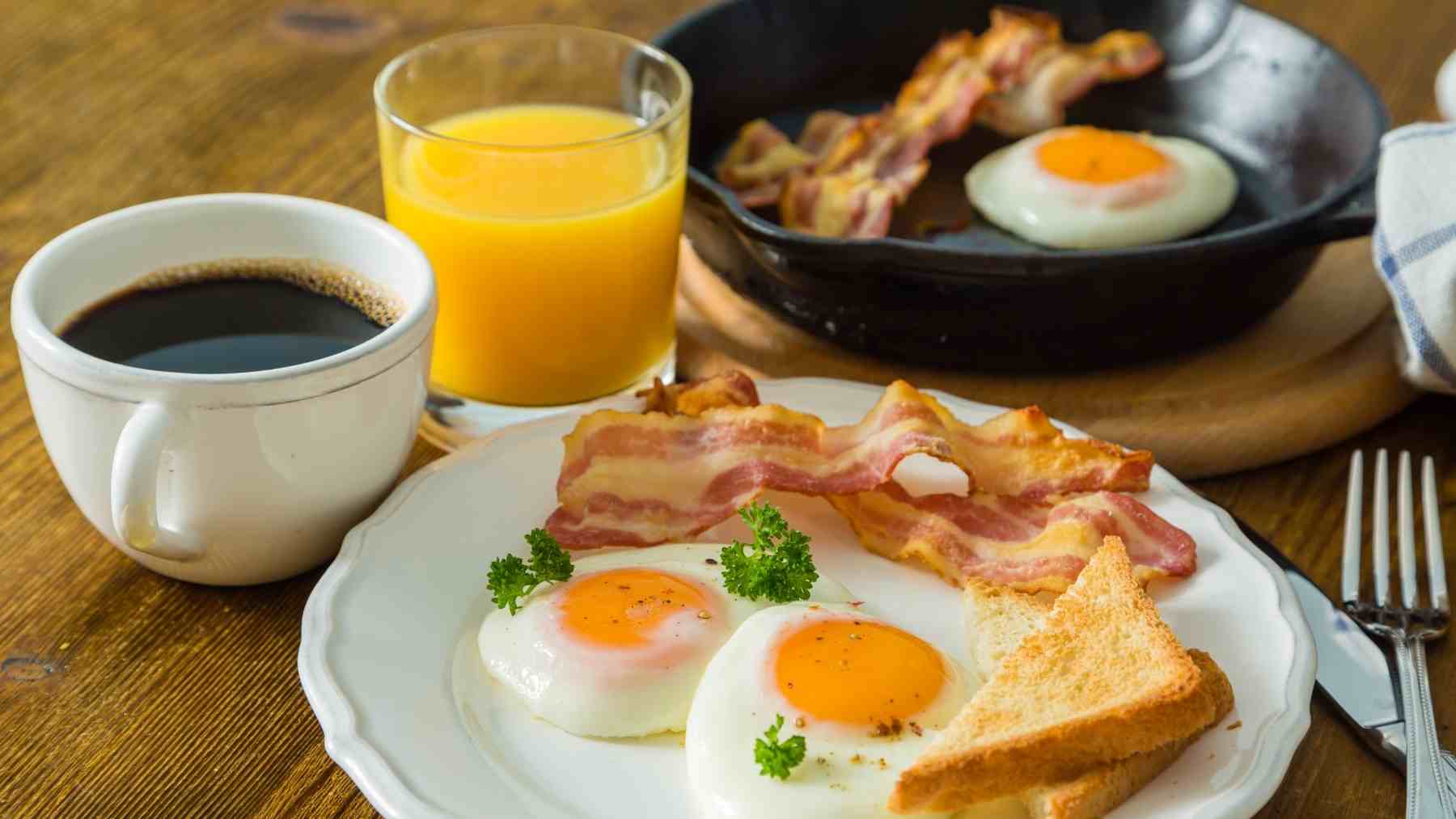 Breakfast with orange juice, black coffee, eggs, bacon, and toast on a wooden table