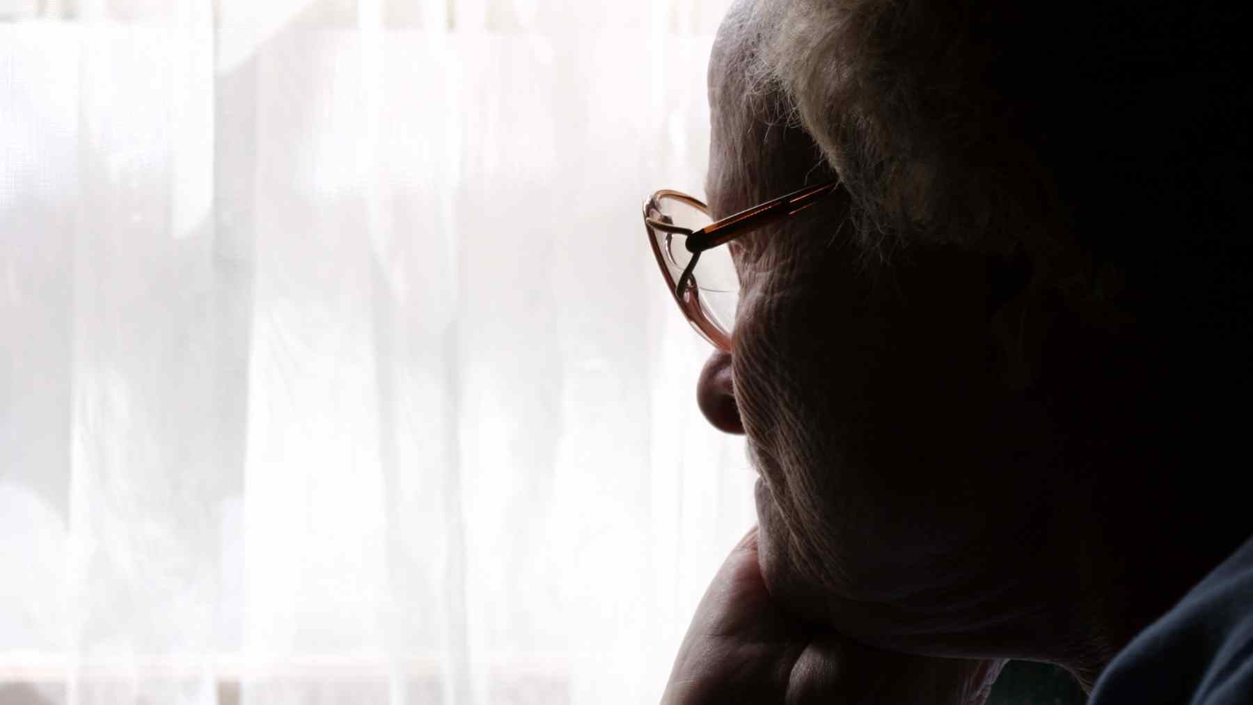 Older parent over 70 sitting in silhouette by a window, reflecting on feeling less needed by adult children.