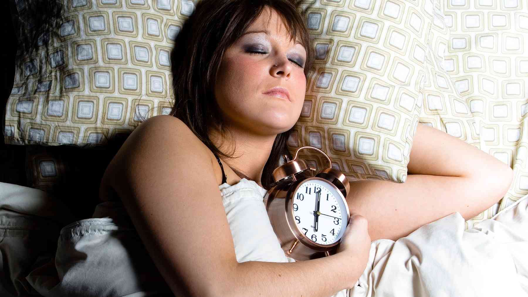 Woman sleeping in bed while holding an alarm clock, illustrating a consistent bedtime and healthy sleep routine.