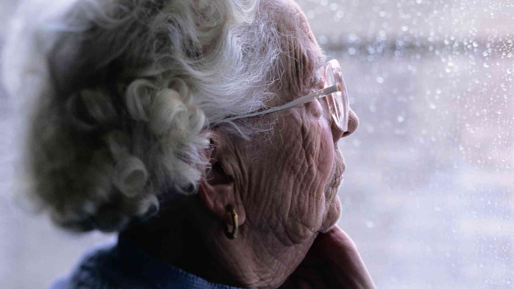 Older woman looking out a rainy window, reflecting the emotional fatigue linked to small talk and feigned interest