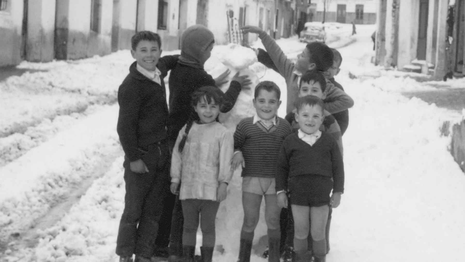 Black-and-white photo of children in a snowy street building a snowman, reflecting independent 1960s-style childhood play