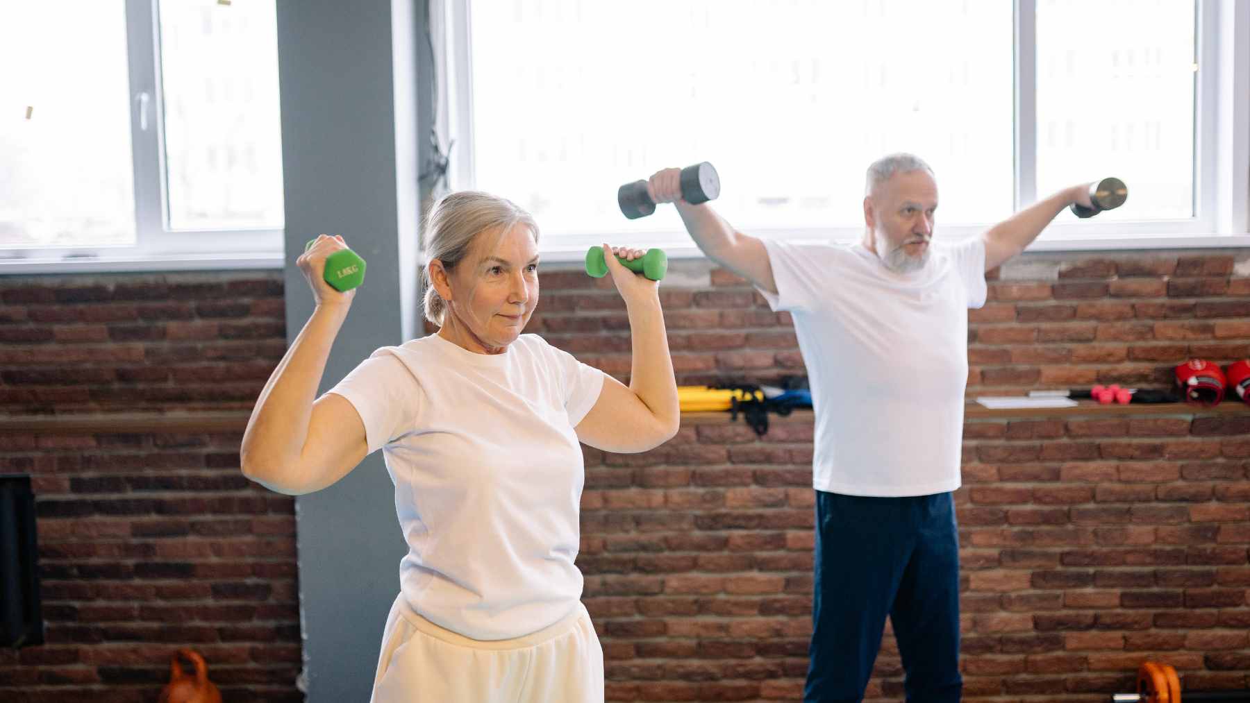 A 76-year-old woman confidently lifting dumbbells during a strength training workout in a bright, modern fitness studio.