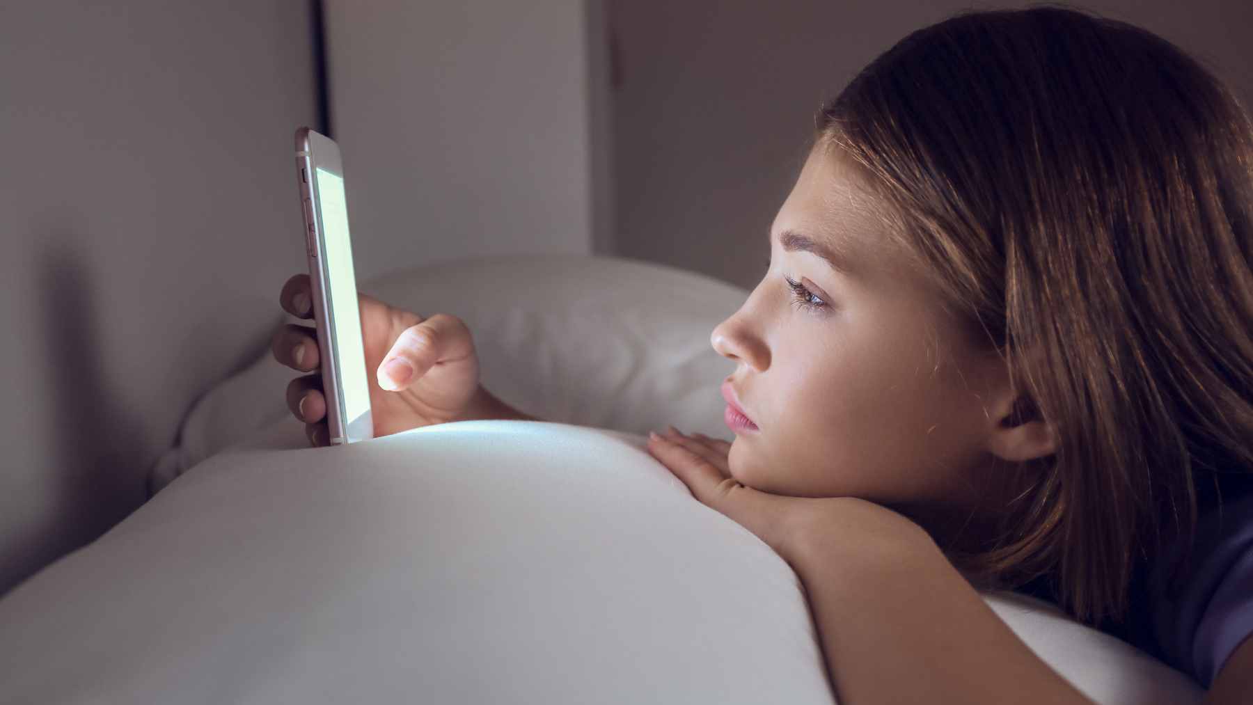 A person lying in bed looking at a glowing smartphone screen in a dark bedroom, illustrating the impact of evening light exposure on sleep.