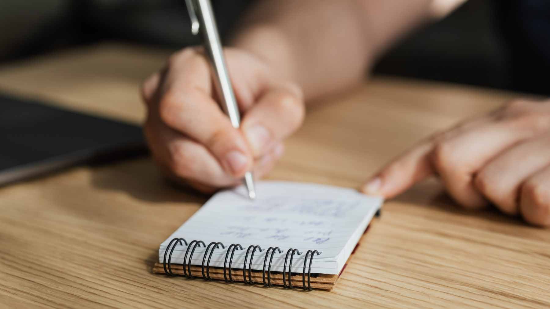 A close-up view of a person taking handwritten meeting notes in a notebook, surrounded by blurred office technology.