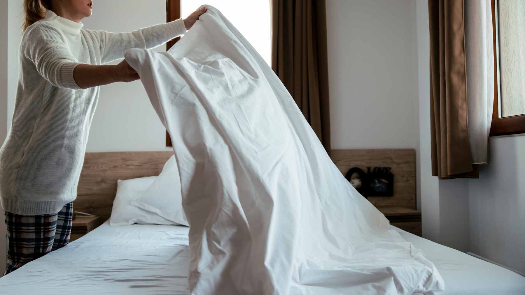 A person tidying their bed covers in a bright, modern bedroom during the early morning.
