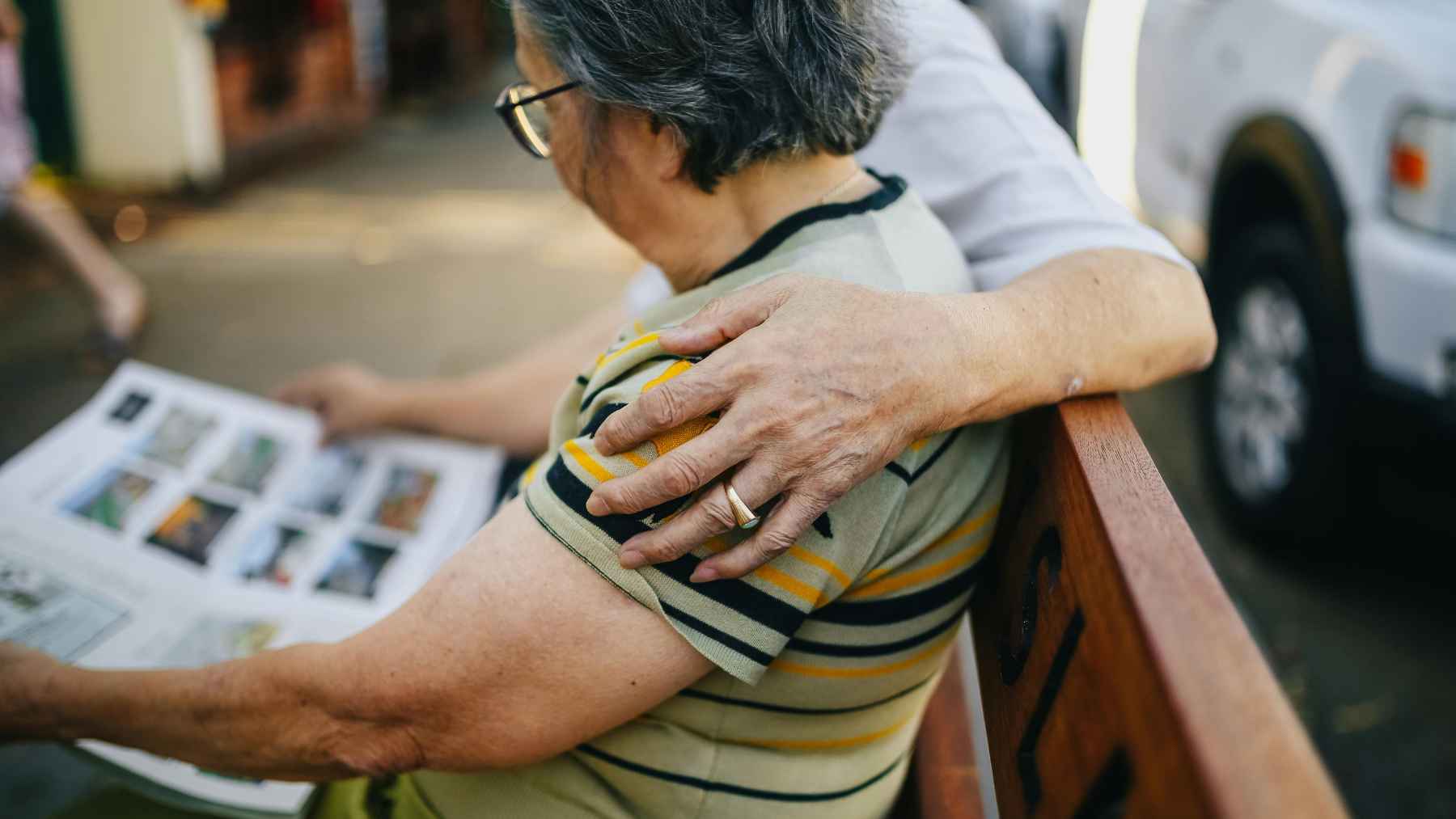 Caregiver comforting an older adult with dementia while looking through printed photos outdoors.