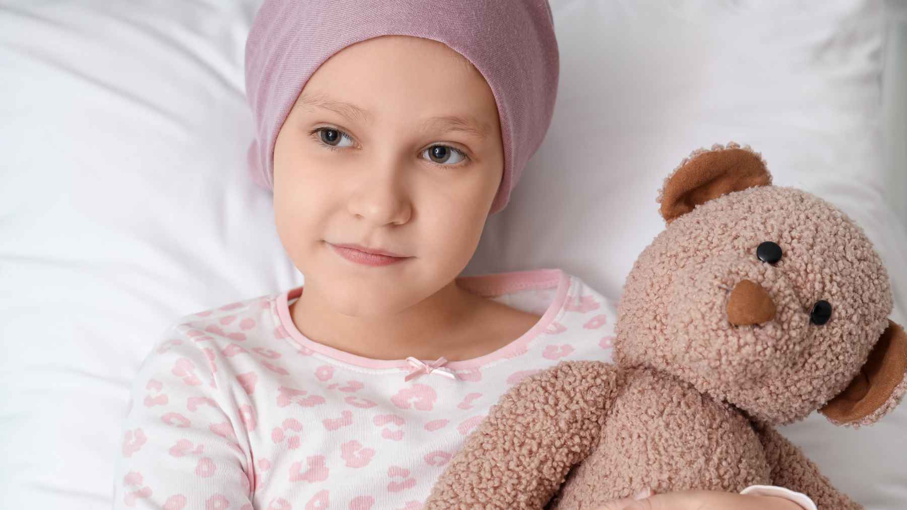 Young child wearing a head covering and holding a teddy bear during pediatric cancer treatment in a hospital room.