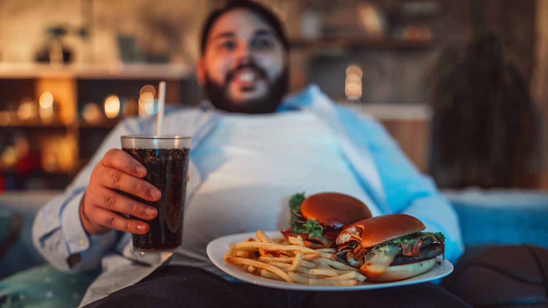 Person holding a sugary soft drink beside burgers and French fries, illustrating an ultra-processed diet associated with chronic inflammation and poor metabolic health.