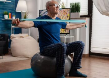 Un jubilado hace ejercicios de fuerza en su sala de estar: sentado sobre una pelota suiza, estira el brazo con una banda elástica de resistencia. Foto: DC Studio / Magnific.