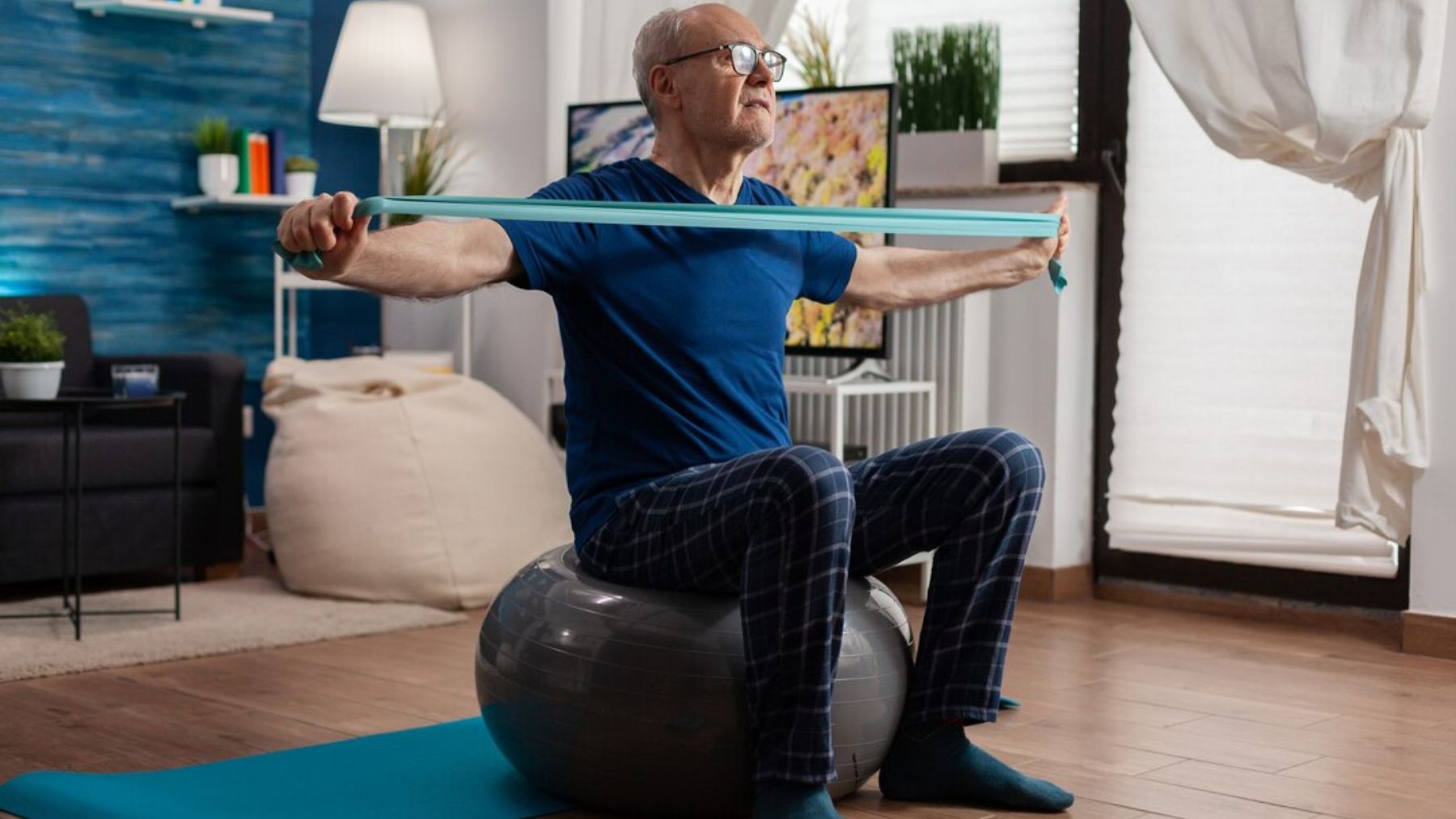 Un jubilado hace ejercicios de fuerza en su sala de estar: sentado sobre una pelota suiza, estira el brazo con una banda elástica de resistencia. Foto: DC Studio / Magnific.