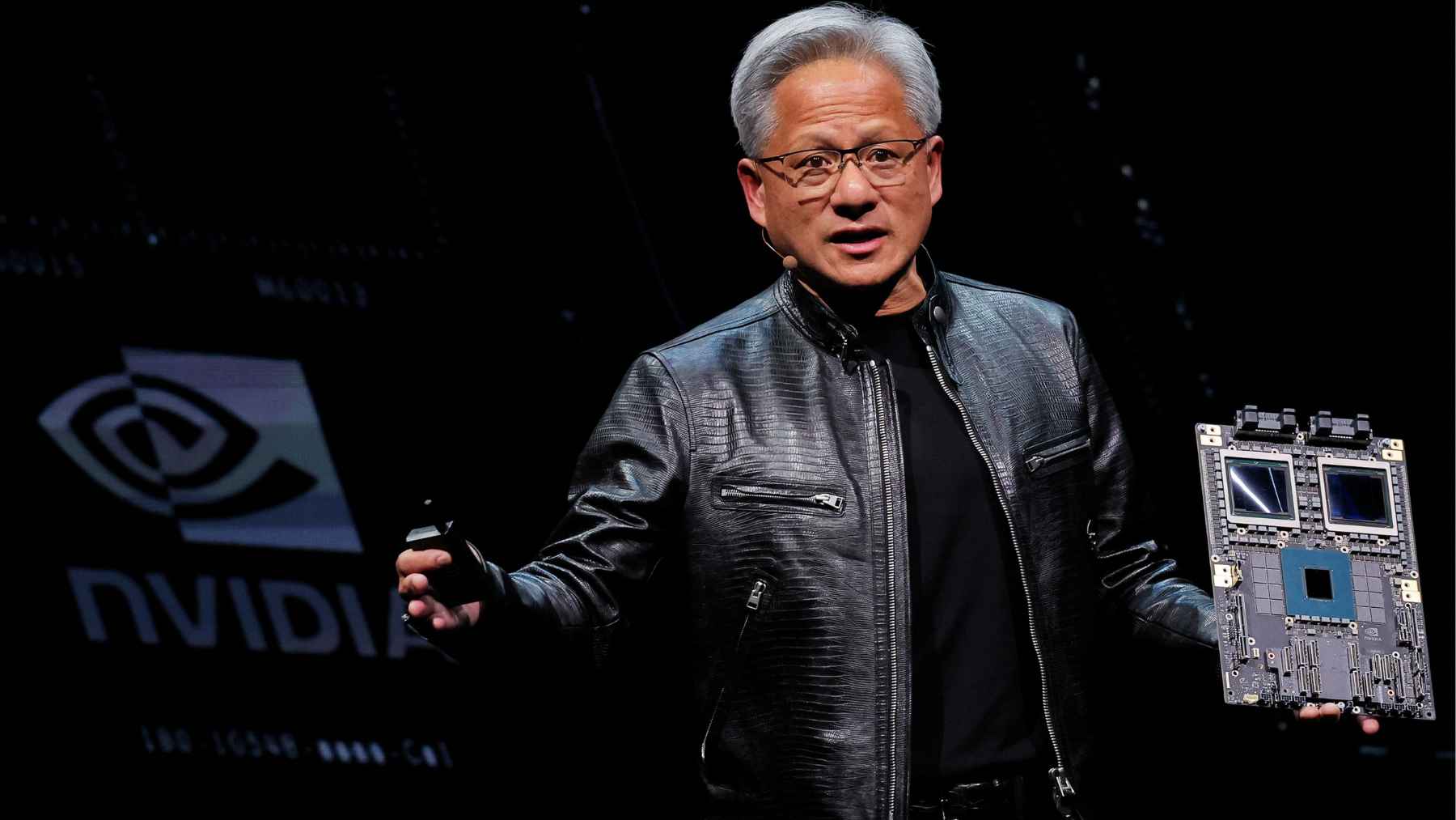 Jensen Huang speaking onstage while holding an Nvidia AI chip board, illustrating the company’s renewed China-compliant chip production