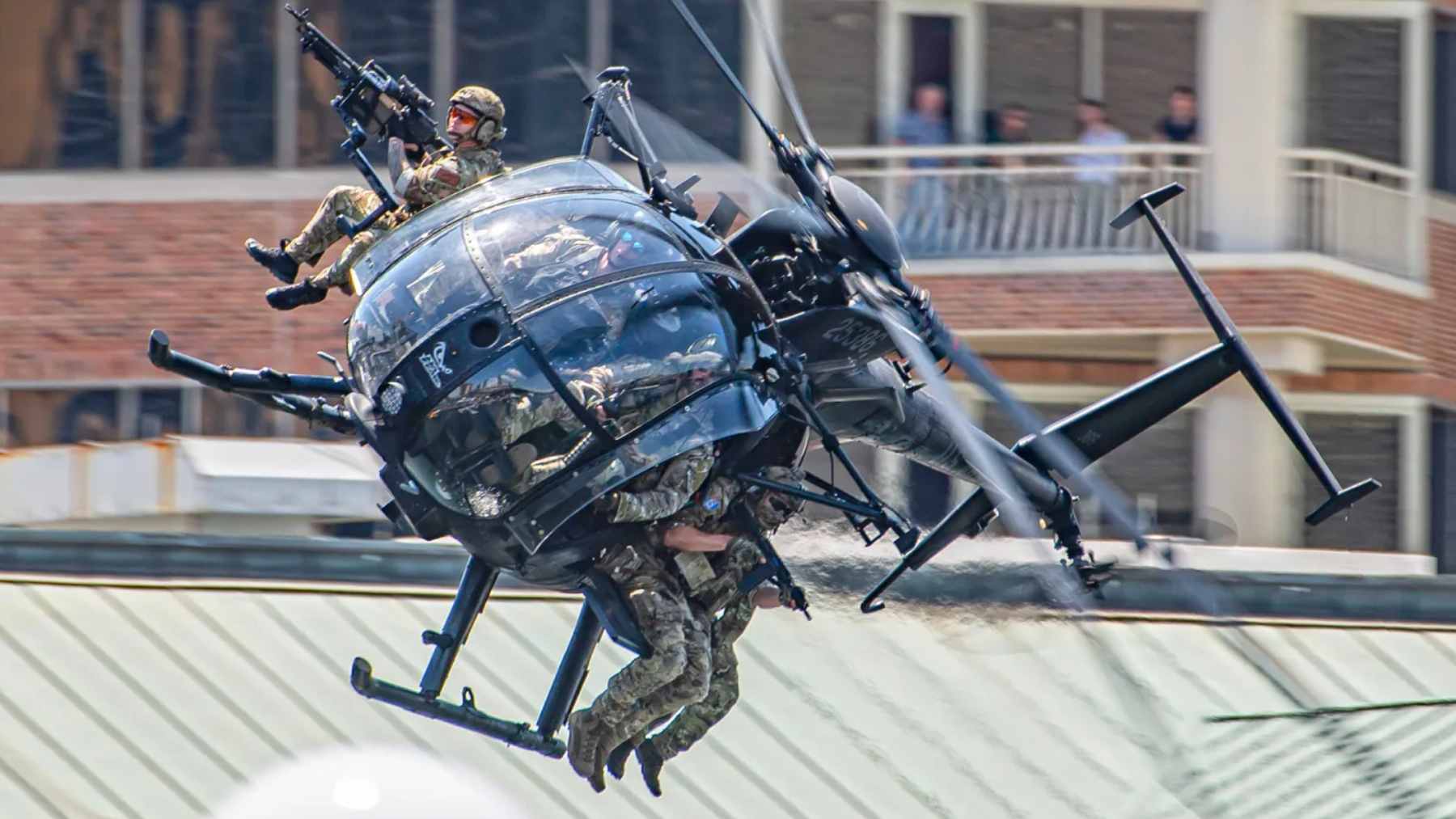 A small, dark MH-6 Little Bird helicopter flying low over rugged, mountainous terrain.