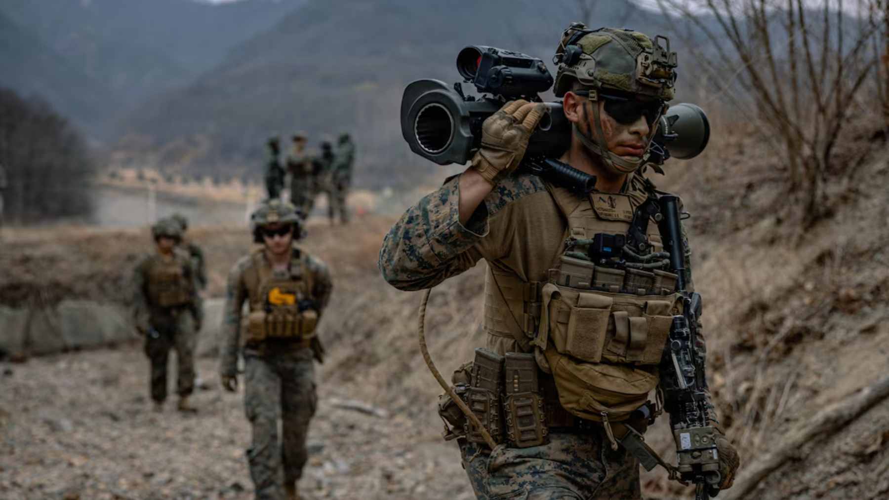 A group of heavily equipped U.S. Marines conducting a search operation in a dense, rain-soaked forest environment.