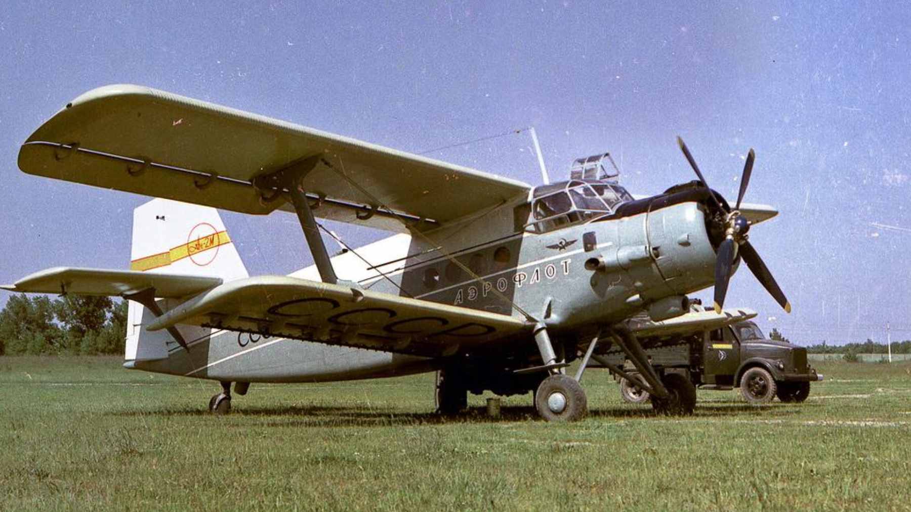 An aging Soviet-era Antonov An-2 biplane parked on a rural grassy airfield.