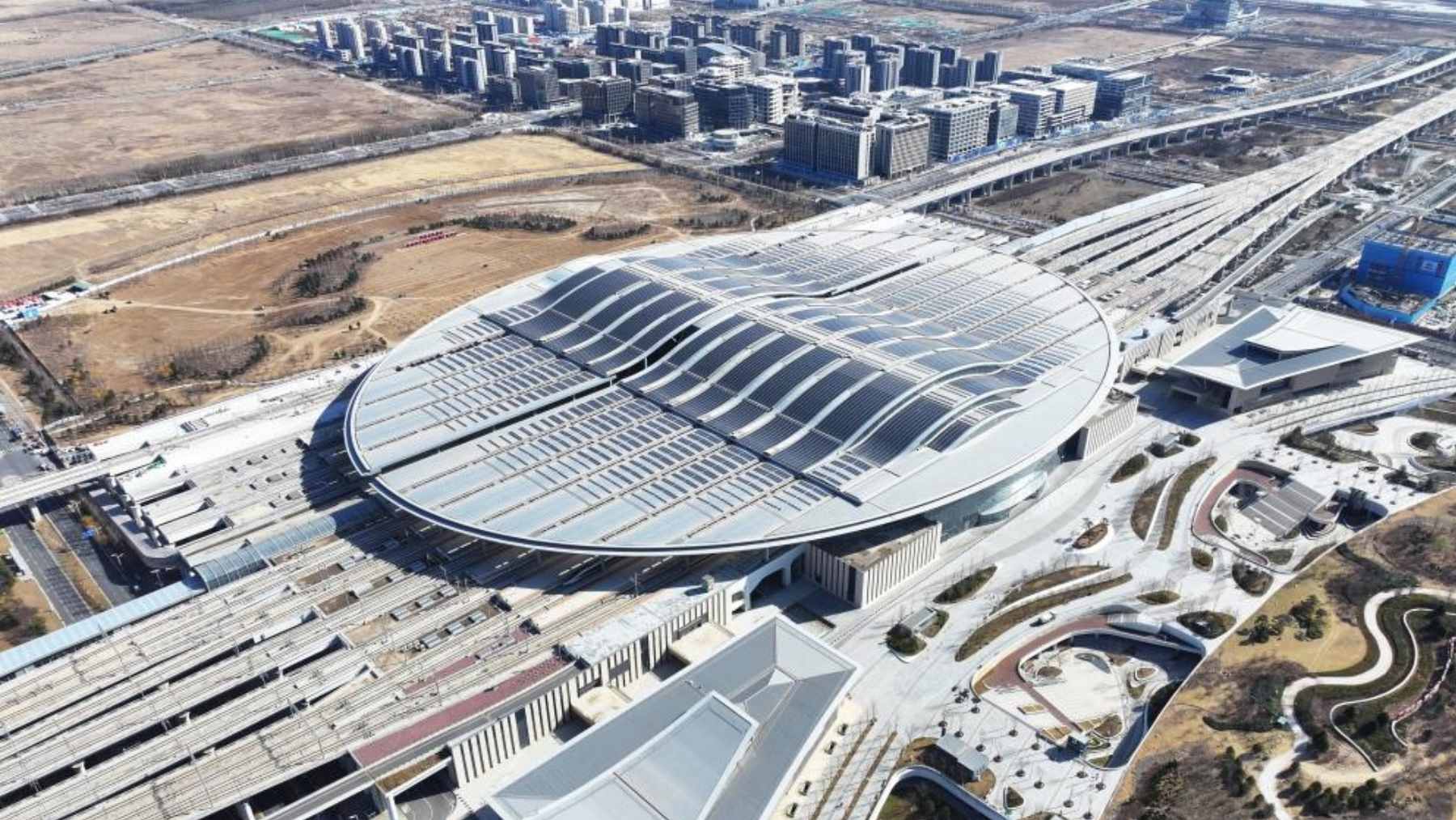 Aerial view of the oval-shaped Xiong’an Railway Station, showcasing its massive roof integrated with solar panels.