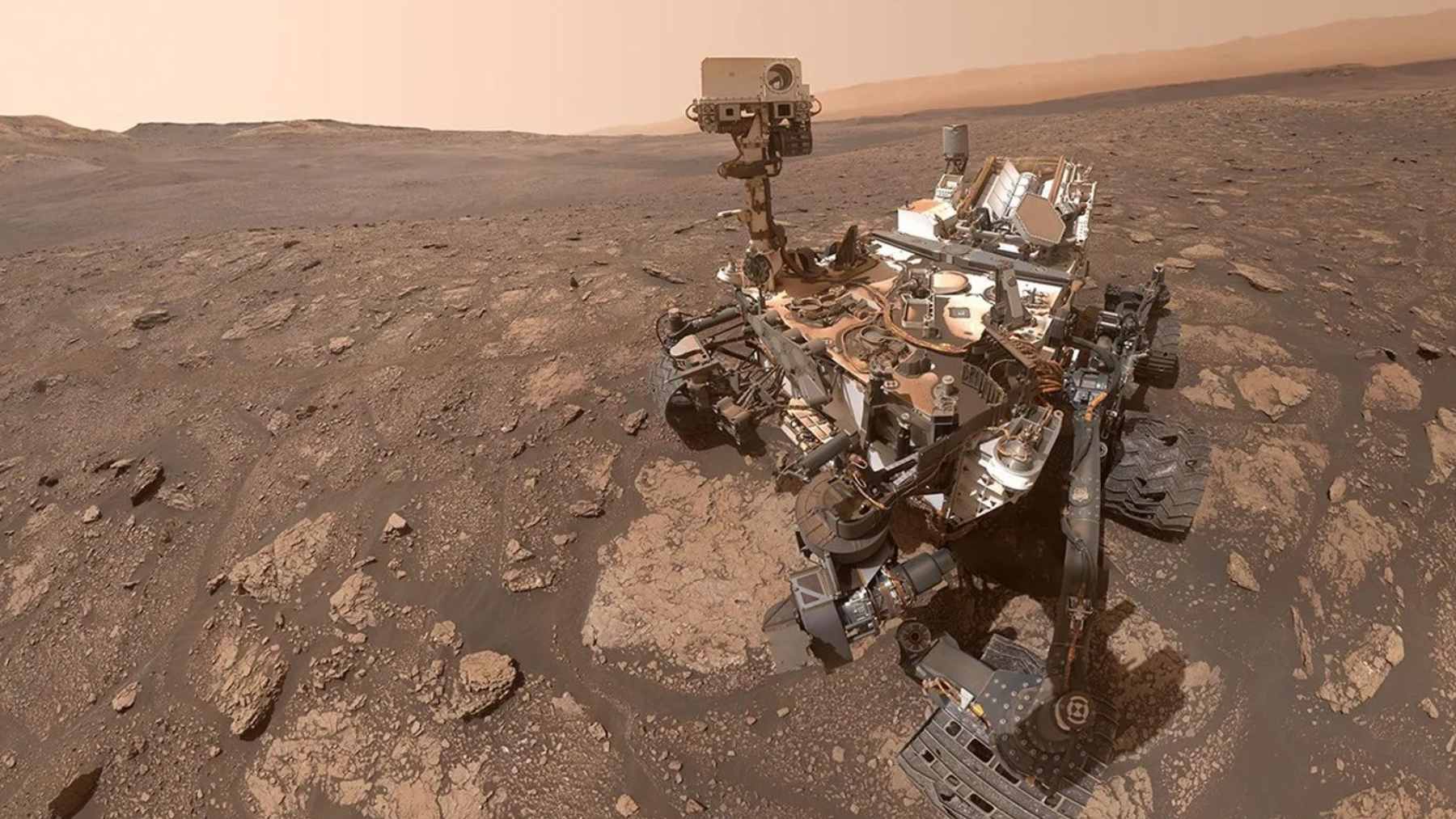 The NASA Curiosity rover parked on the Martian surface with the layered slopes of Mount Sharp visible in the background.