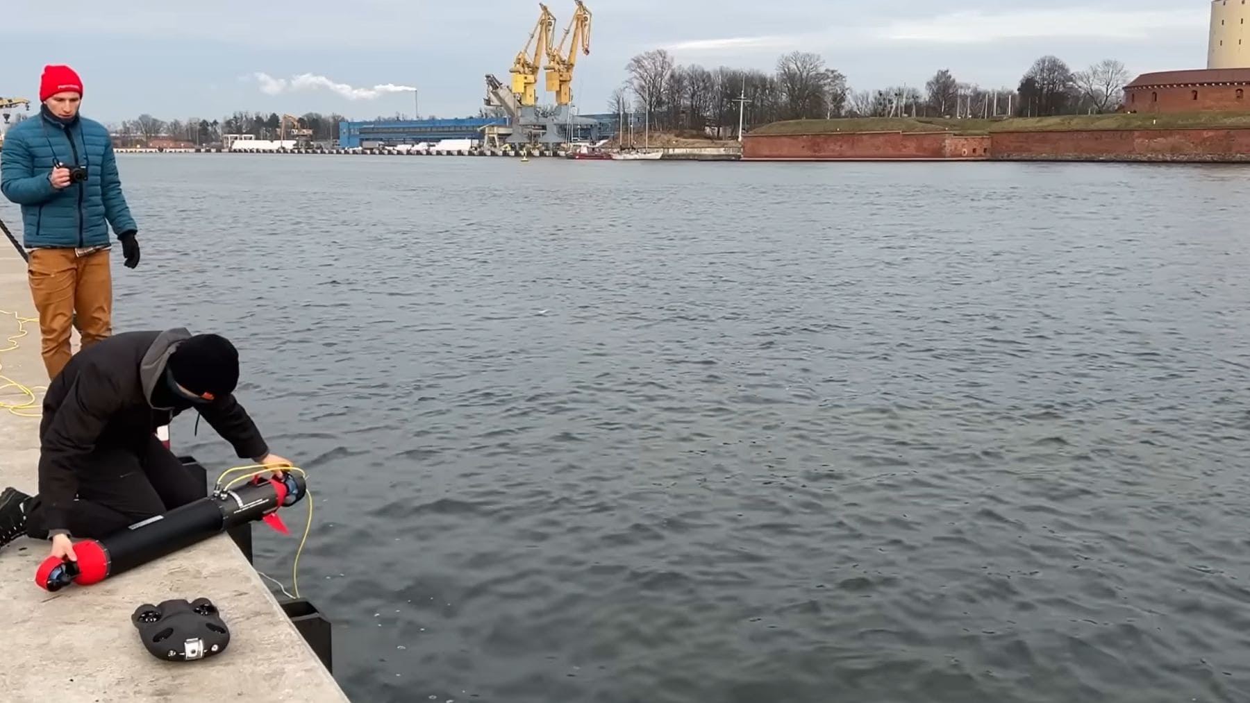 Un joven con gorro rojo opera un dron submarino casero tubular negro con aletas rojas desde un muelle de cemento, preparándolo para una misión autónoma en un río con corriente.